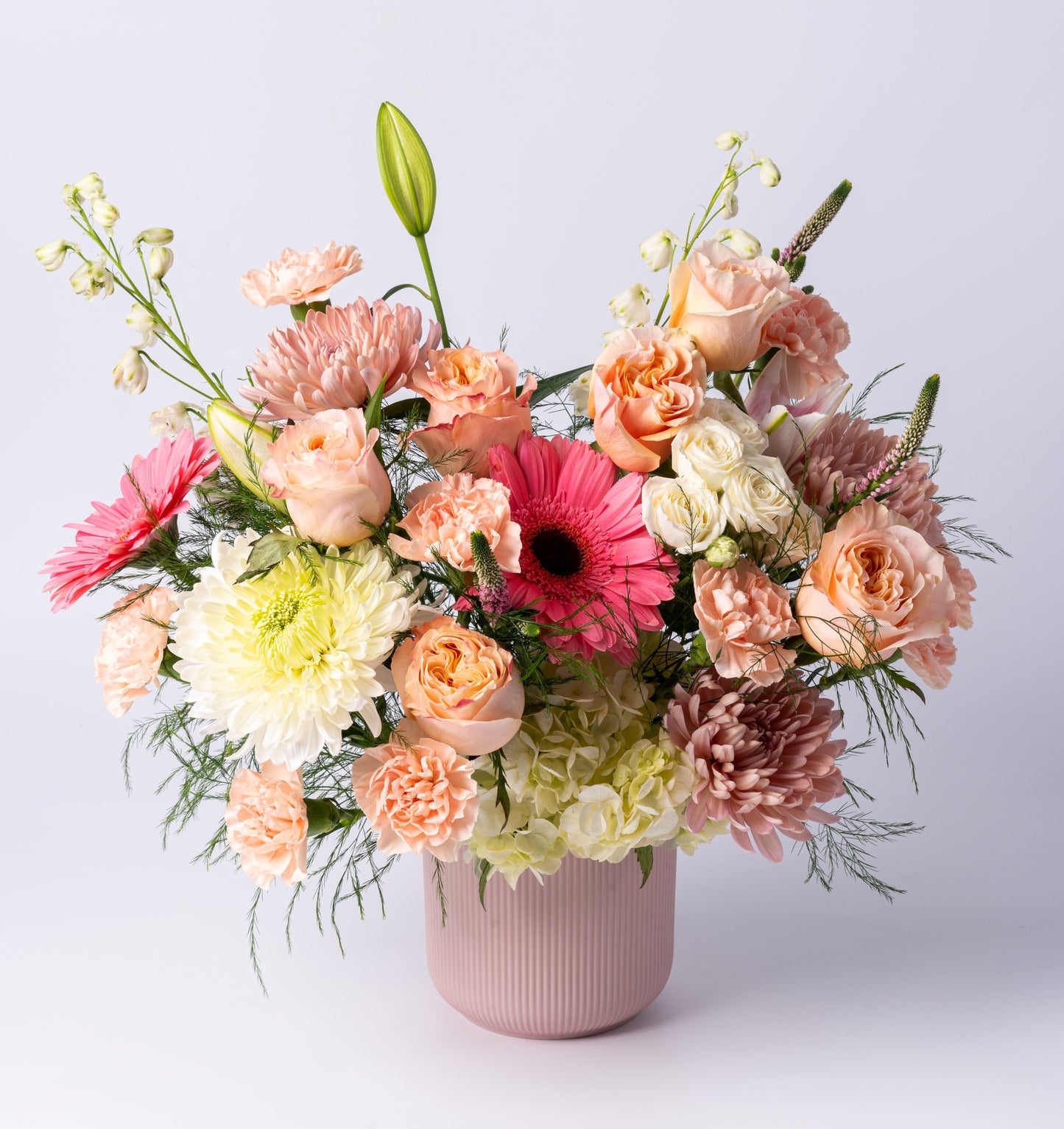 A romantic pastel arrangement with peach roses, gerberas, hydrangea, and blush blooms in a matte pink vase. Soft, elegant, and beautifully lush.