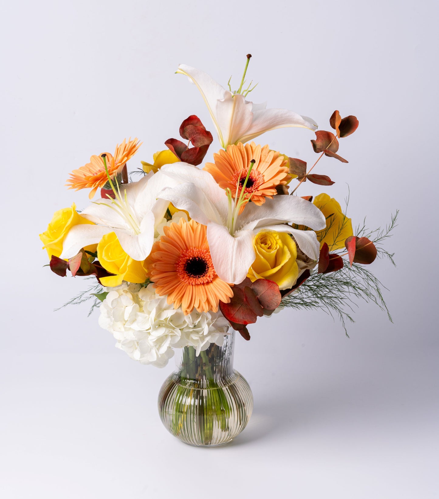 Bright vase arrangement with white lilies, peach gerbera daisies, yellow roses, hydrangea, and bronze eucalyptus in a ribbed glass vase.