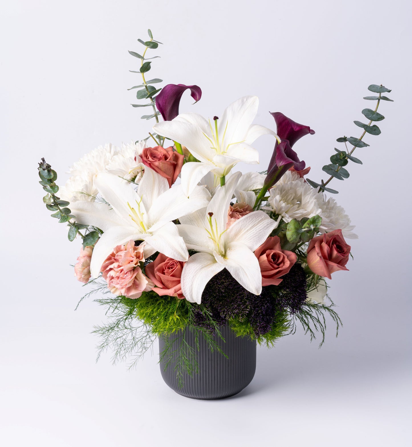 Elegant vase arrangement with white lilies, dark purple calla lilies, blush roses, carnations, chrysanthemums, and eucalyptus in a charcoal ceramic vase