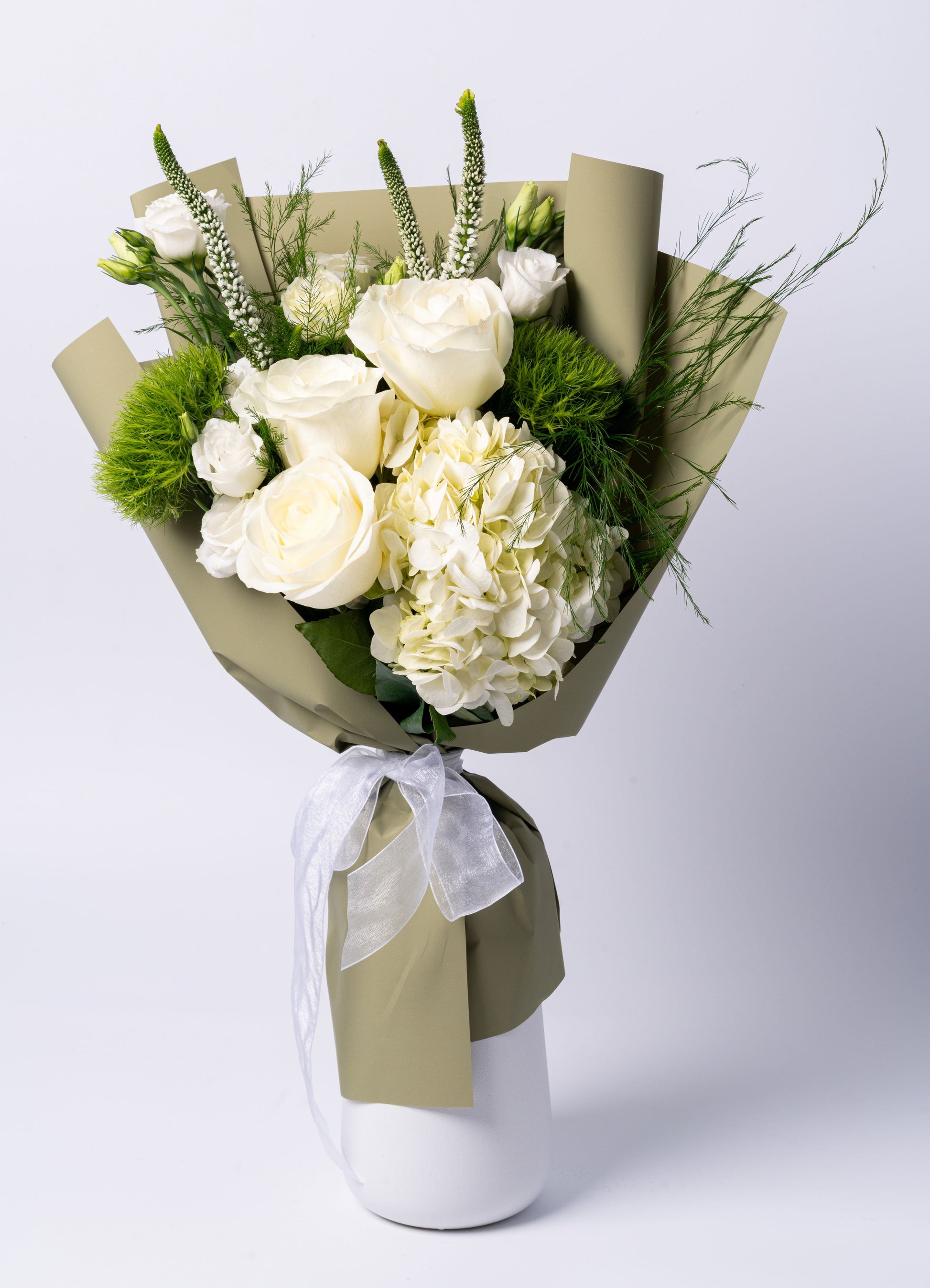 Wrapped bouquet with white roses, hydrangea, lisianthus, veronica, and green moss balls in sage wrapping with a sheer ribbon. Vase not included