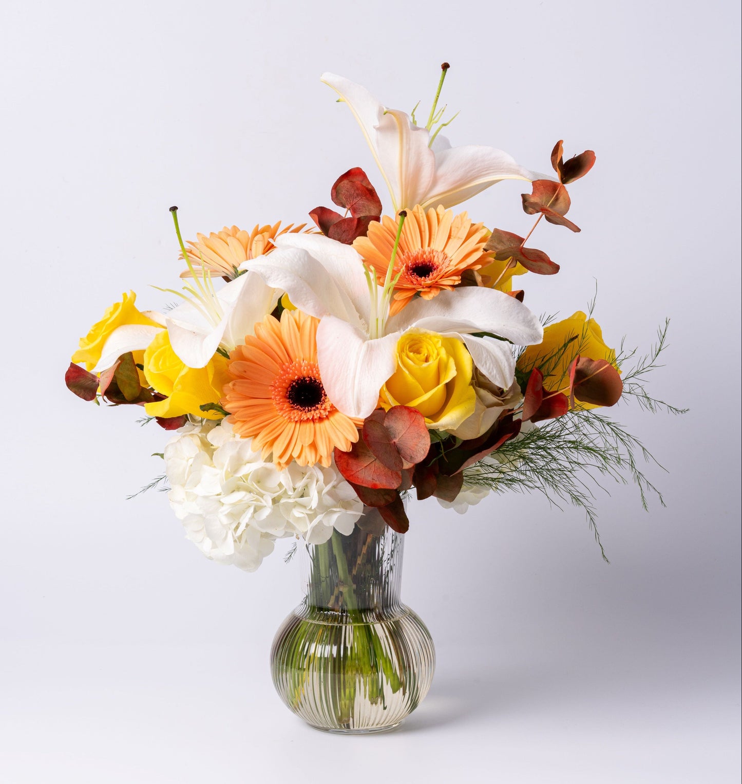 Bright vase arrangement with white lilies, peach gerbera daisies, yellow roses, hydrangea, and bronze eucalyptus in a ribbed glass vase.